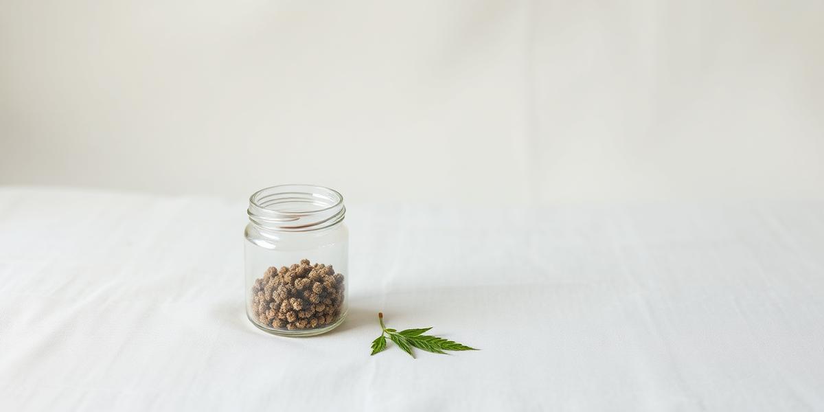 A clear glass jar of dried hemp flower next to a single hemp leaf on pale linen